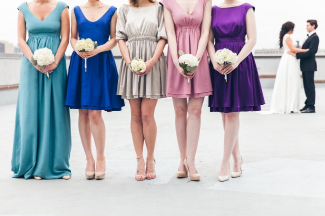 Bridesmaids wearing durga-kali dresses in pink, blue, teal, purple and grey colors holding white flower bouquets. bride and groom in the back ground