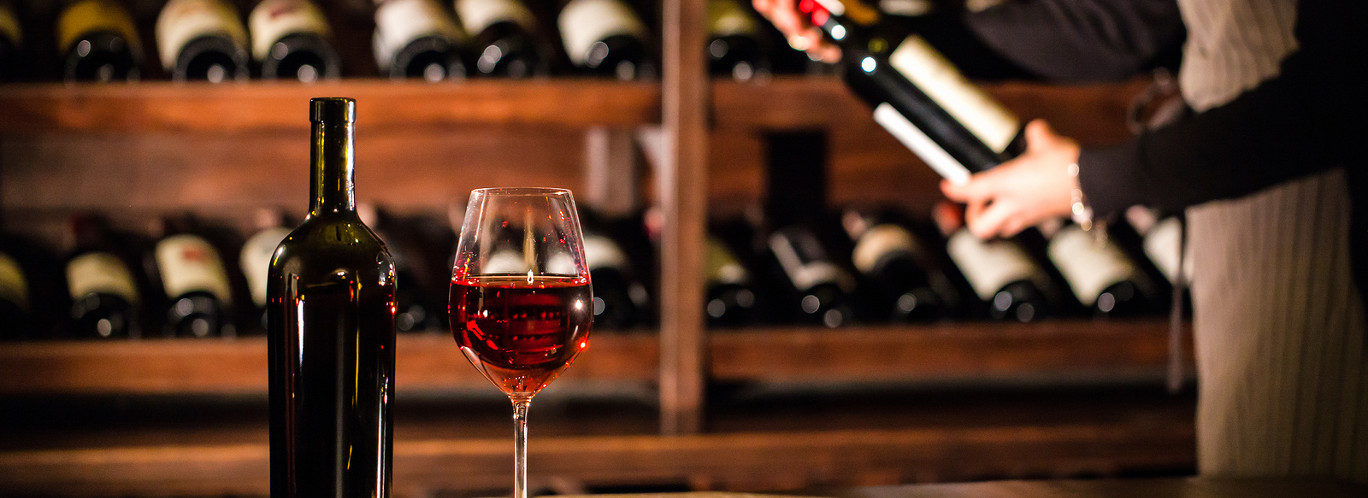 Glass of red wine and a bottle place on a table. Sommelier holding a bottle of wine and standing next to the shelves with wine bottles.