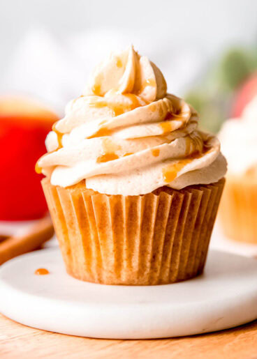 an apple cinnamon cupcake topped with caramel buttercream and caramel sauce on a white plate; background has cinnamon sticks and red apples