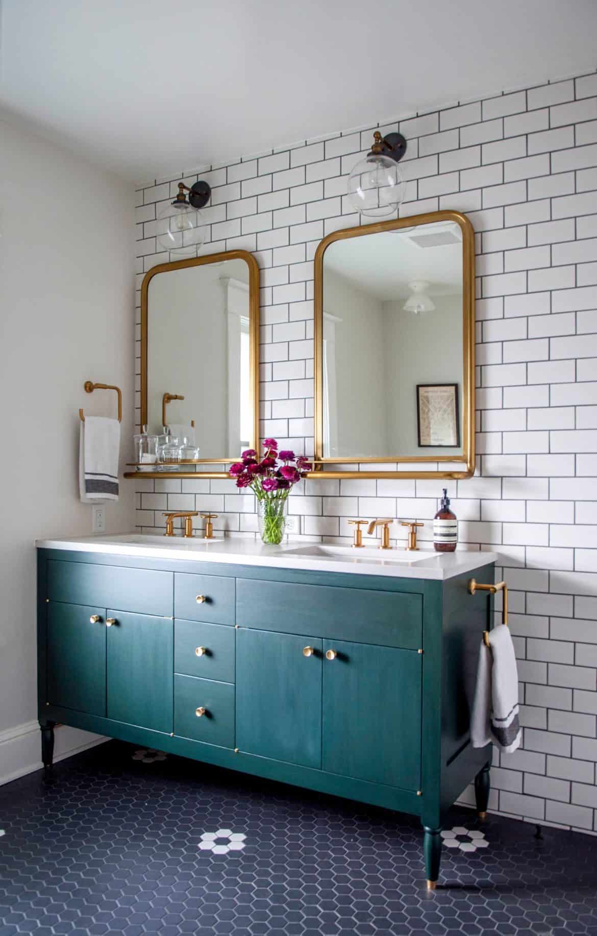 This bold and colorful vintage modern bathroom  has one wall covered completely with white subway tiles. Two gold-framed mirrors hang over a double vanity, with hunter green painted cabinets and a white marble countertop. The bathroom floor is tiled with navy blue hexagonal tiles.