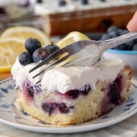 a fork cutting into a piece of lemon blueberry cake on a white plate with fresh blueberries on top