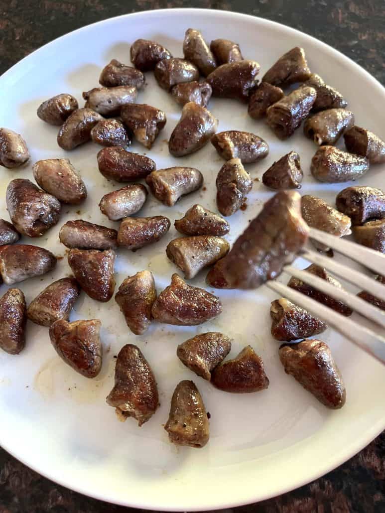 Fork holding a pan-fried chicken heart over a plate filled with golden-brown cooked chicken hearts, ready to be enjoyed.