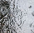 A puddle on a gravelled surface with raindrops hitting it, creating a pattern of concentric circular ripples on its surface.