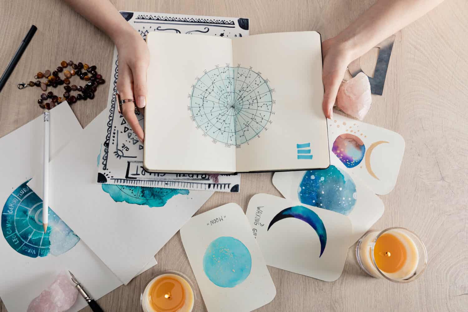 Top view of astrologer holding notebook with watercolor drawings and zodiac signs on cards on table