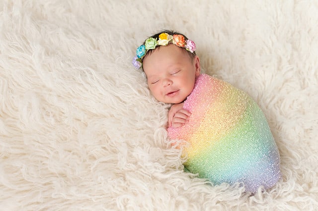 A smiling nine day old newborn baby girl bundled up in a rainbow colored swaddle. She is lying on a cream colored flokati (sheepskin) rug and wearing a crown made of roses.