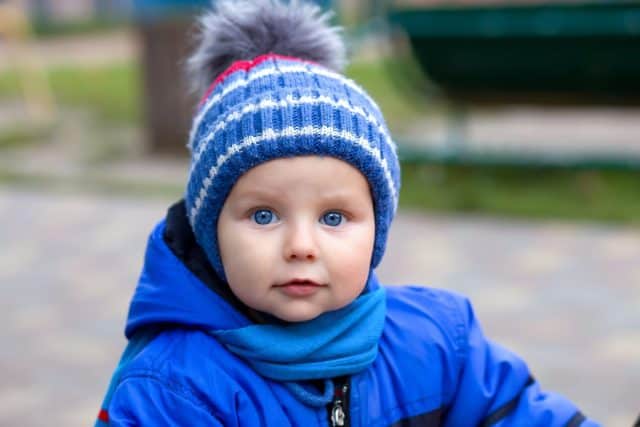 A one-year-old blue-eyed baby in a blue overalls and a hat looks into the frame and smiles. baby learning to walk holding mother's hand