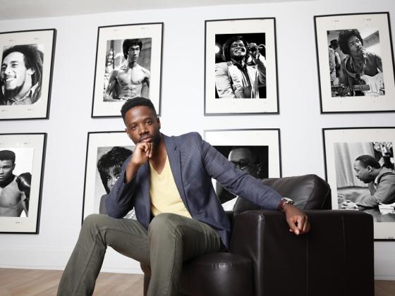 A man in a sportcoat sits in a chair with photos of iconic figures behind him.  | 