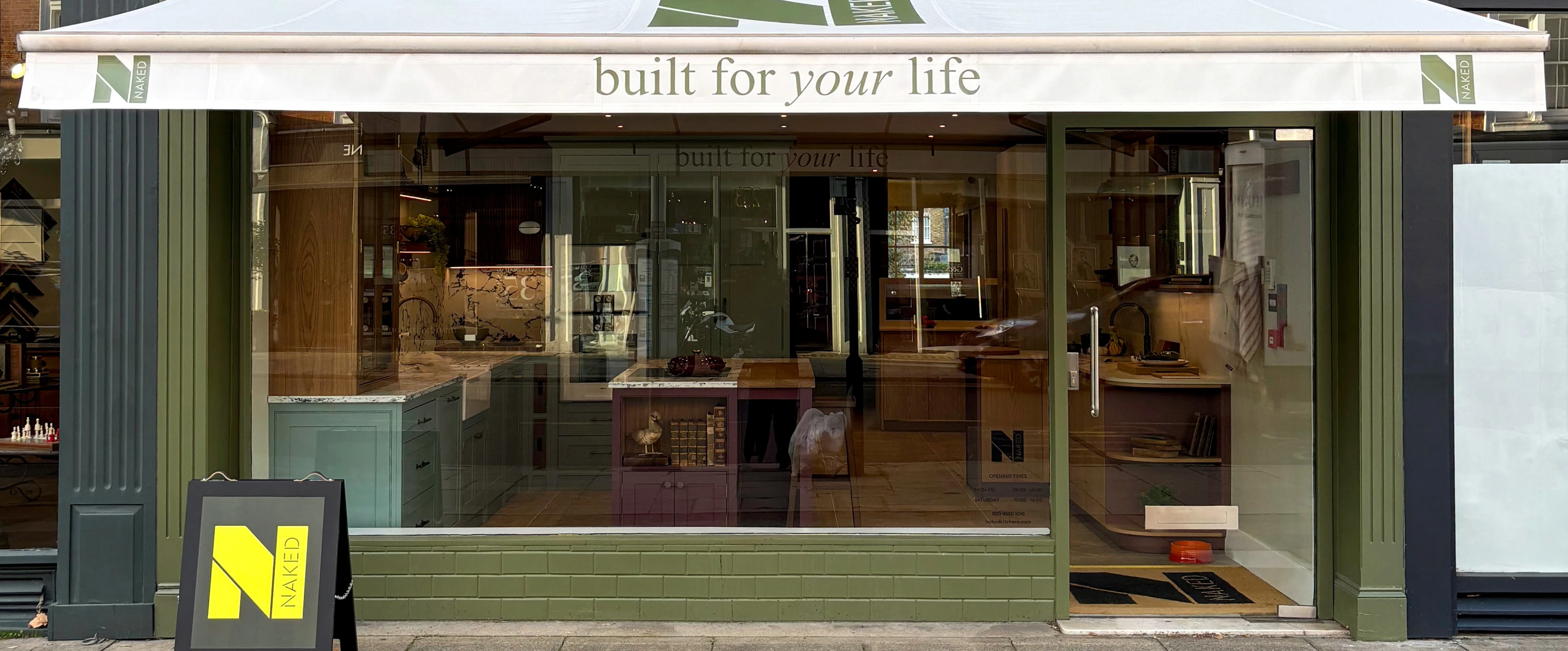 Street view of Naked Kitchens storefront with a green facade and glass door. An awning displays 'built for your life'. A small sign with the logo and name stands on the sidewalk. The interior shows a modern kitchen display.