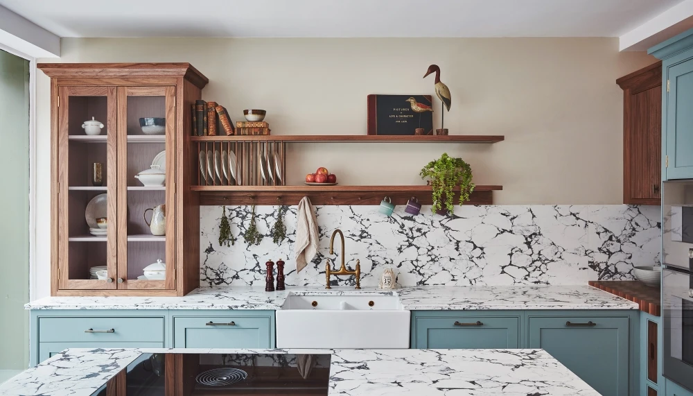 A kitchen with blue cabinets, marble countertops, and a farmhouse sink. Wooden shelves hold dishes, plants, and decorative items. A tall cabinet displays more dishes. A backsplash with a black and white pattern complements the design.