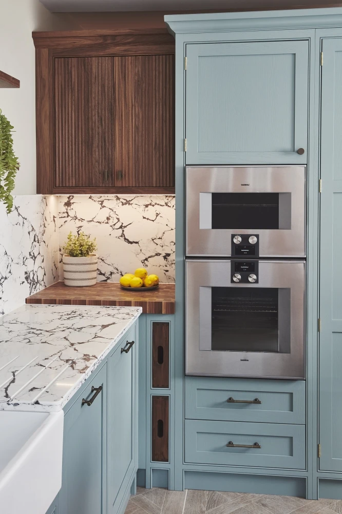 Modern kitchen with a light blue cabinet featuring built-in stainless steel ovens. The countertop and backsplash have a white and gray marble pattern. A small wooden section holds lemons, a plant, and features an underlit dark wooden cabinet.