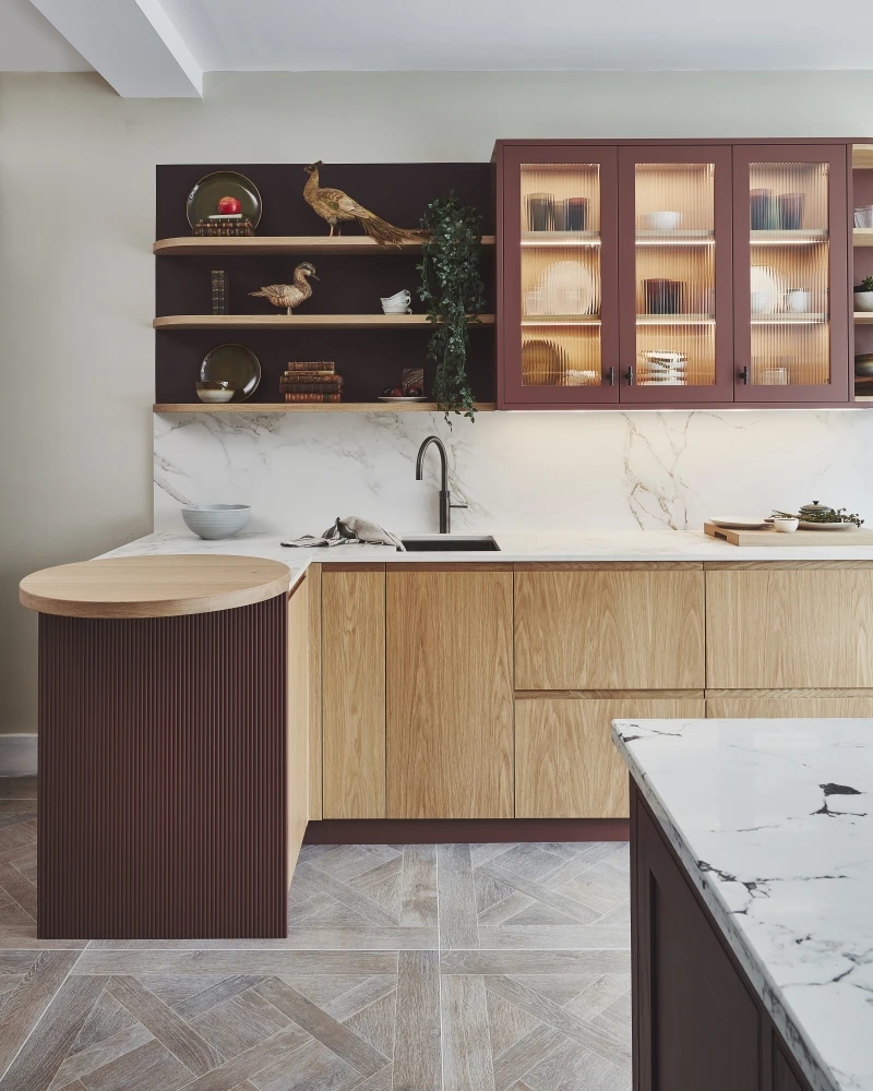 Modern kitchen with wooden cabinets, marble countertops, and an island. Cabinets display decorative items like a pheasant, plates, and a plant. The floor features a herringbone pattern. A sink is installed under mounted shelves.