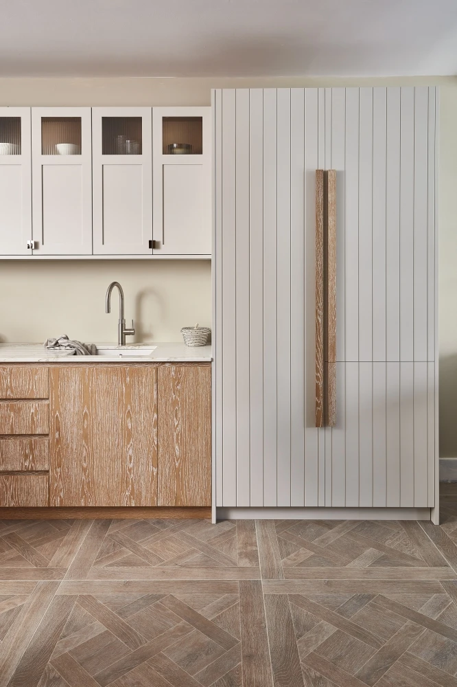 A modern kitchen features light wood cabinets, a white vertical-paneled refrigerator, and upper cabinets with glass inserts. A stainless steel faucet is positioned over a sink on a stone countertop. The floor is patterned wood.