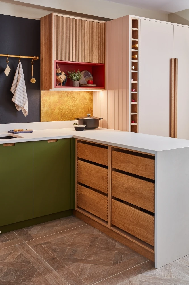 Modern kitchen with a white countertop and wooden cabinets. The design features green lower cabinets, vertical wine storage, and a black backsplash with hanging hooks. Decorative items and a potted plant are on the countertop. Geometric tile flooring.