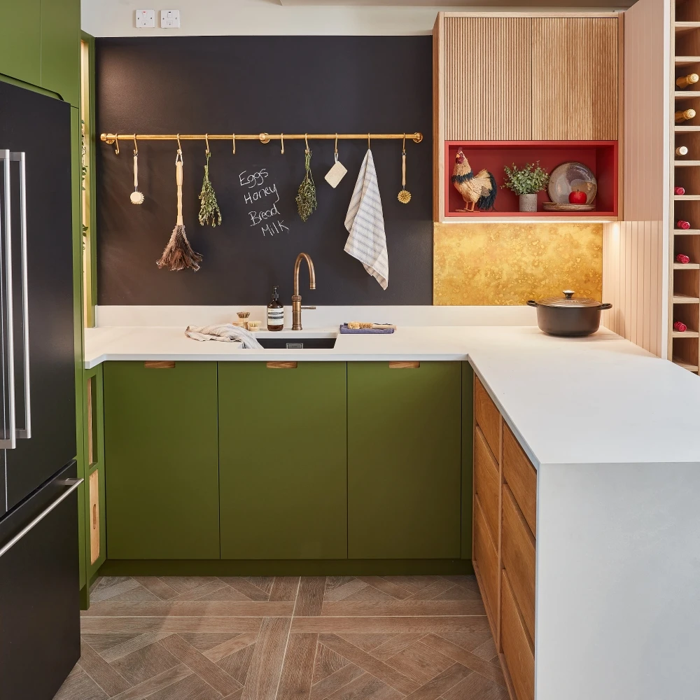 Modern kitchen with green and wood cabinets, blackboard wall with shopping list, and decorative shelf. Features a gold faucet, white countertop, and wine rack. Hanging utensils and a pot on the stove add functional touches.