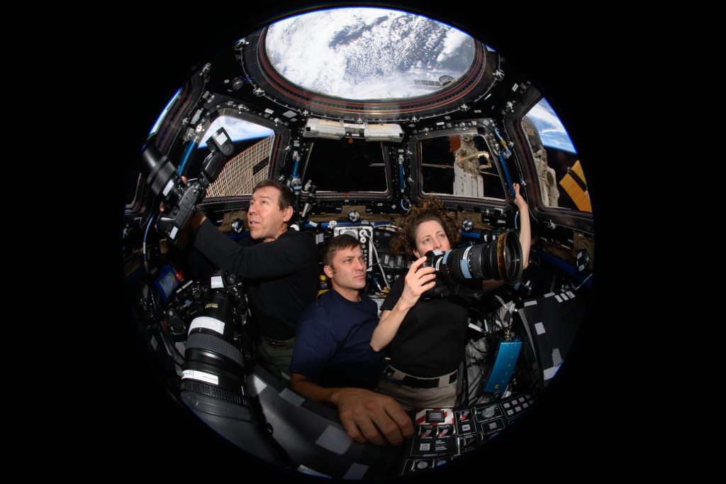 Expedition 70 Flight Engineers (from left) Mike Barratt, Matthew Dominick, and Loral O'Hara take photos of the Earth from inside the cupola. Barratt and O’Hara hold large lens cameras, pointing towards the cupola window facing the Earth. Dominick floats behind them also looking up at cupola window. The photo is taken with a fisheye lense causing the photo a circular appearance.