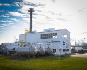 A side view of a white building with a tall metal pole sticking out of the top, large metal cylinder equipment next to it, and a blue sky with clouds in the background.