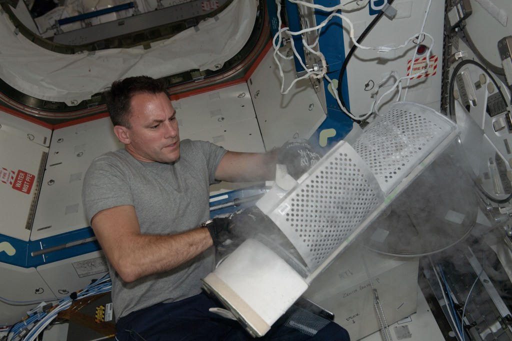 An astronaut wearing a gray shirt and gloves handles a frosty, smoke-emitting component inside the International Space Station.