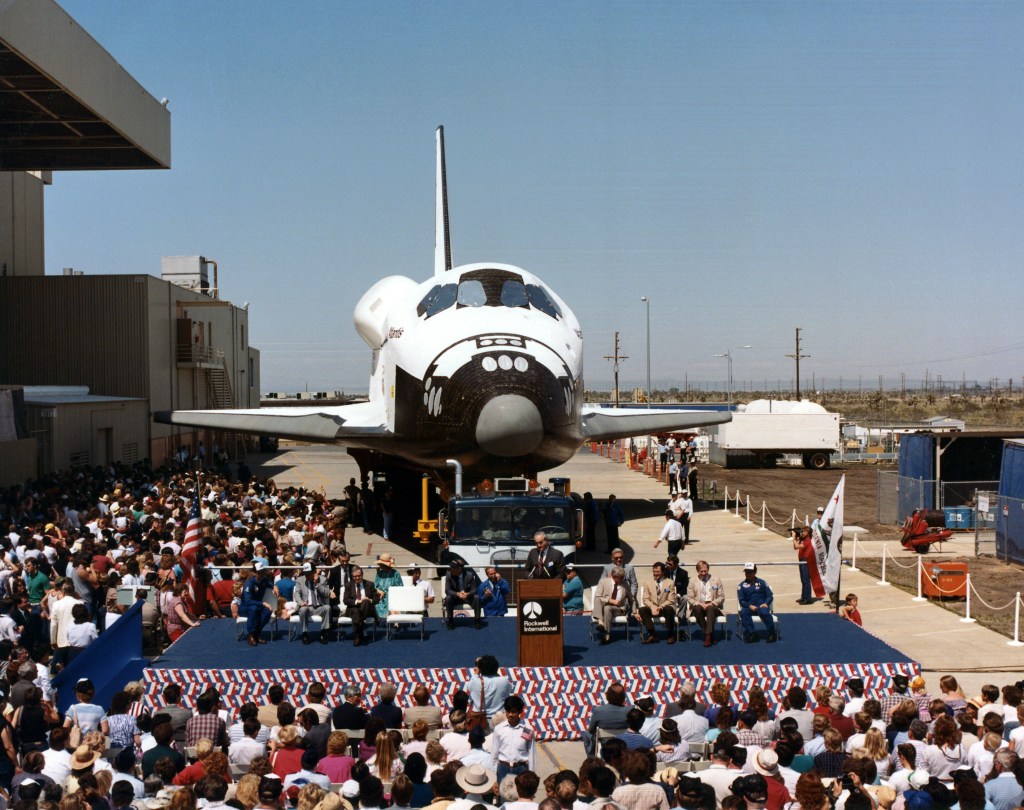 View of a space shuttle orbiter with a about a dozen people on a stage in front of it and an assembled crowd.