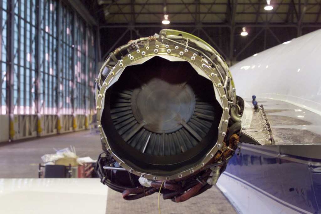 A close-up of a chevron nozzle with jagged edges installed on the engine of a white aircraft with a blue stripe along its side. The aircraft is inside a large hangar.