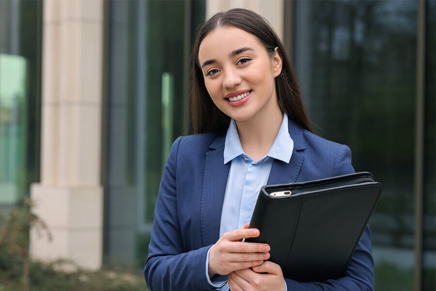 Smiling businesswoman in a blue suit holding a folder outside an office building.