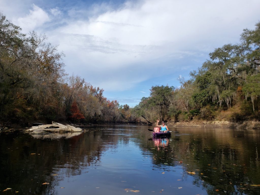 Woman in purple kayak outdoors waving from middle of the river