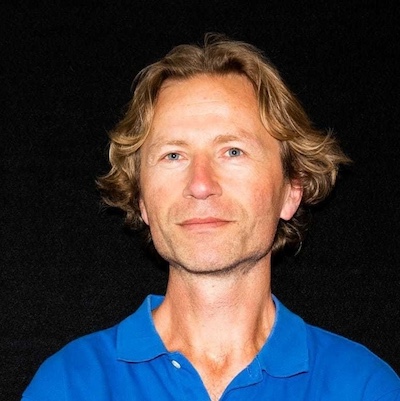 A man in a blue polo shirt standing against a black background.