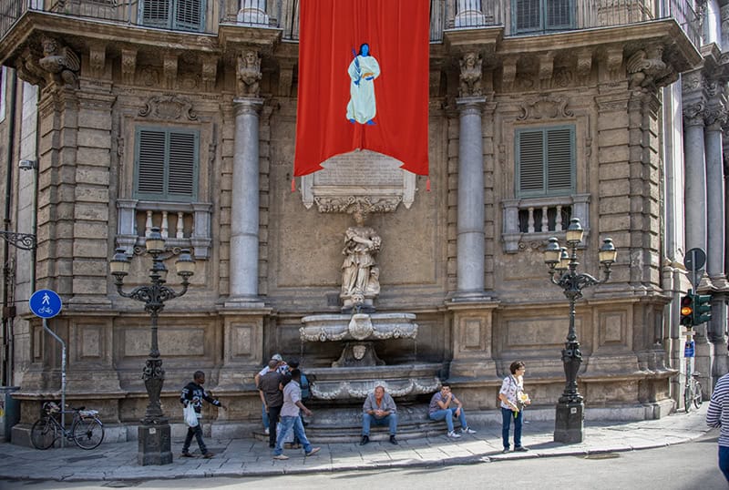 men sitting below a large red flag on a street corner