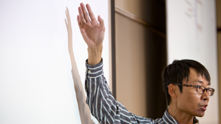 A side profile image of an asian man standing in front of a whileboard teaching a class.