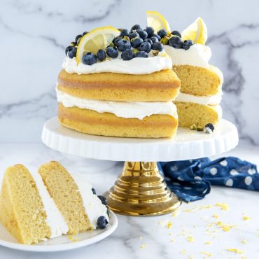 2 Layered cake with frosting in between layers and on top with fresh blueberries and lemon slices, one cut piece.