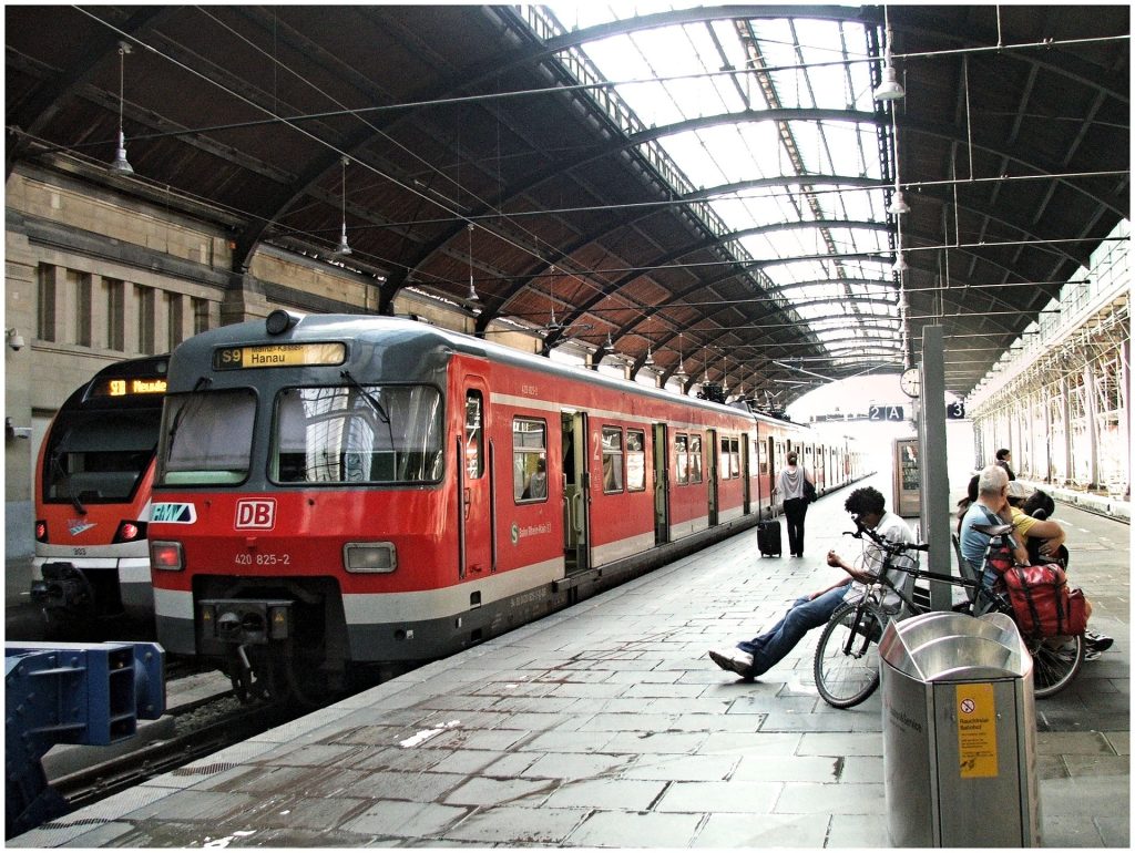 Bahnsteig 2A eines Bahnhofs, wo ein roter DB Zug steht mit DB Logo vorne. Der Zug fährt nach Hanau. Am Bahnsteig sitzen Personen die warten und eine Person geht mit dem Koffer auf den Zug zu.