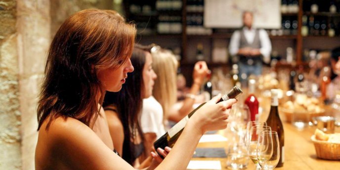 A young woman sits at a long table set with wine glasses while examining a wine bottle during a Paris Wine Cellar Tasting