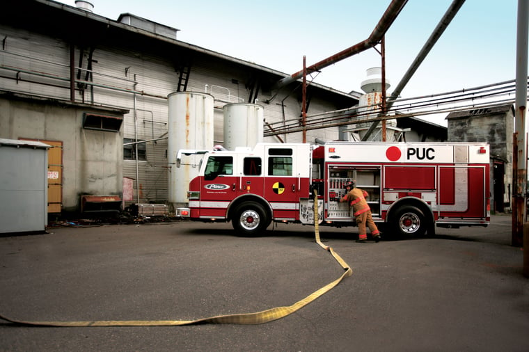 Pierce PUC firetruck parked outside near a building with a firefighter storing equipment in driver’s side storage compartments. 