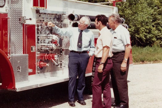 Three men standing next to a Pierce Manufacturing Fire Truck parked outside inspecting the pump panel. 