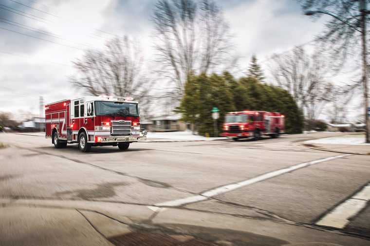 Pierce fire apparatus driving down the road passing a Pierce fire apparatus stopped at a stop sign on a cloudy day. 