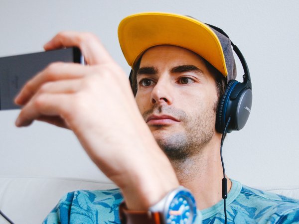 A man wearing a straight-brimmed baseball cap, watching TV on his phone while wearing black over-the-ear headphones.