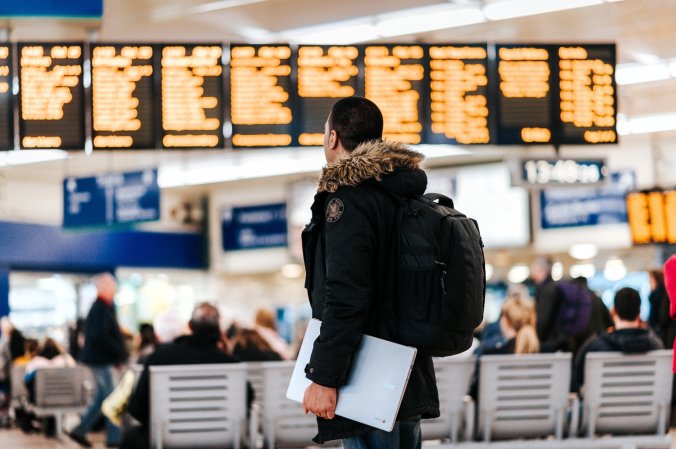 A man holding a laptop in an airport and looking at the flight board.