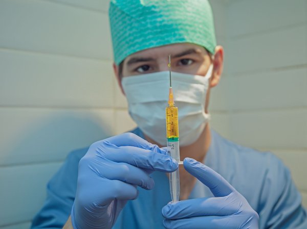 A doctor wearing a mask and scrubs holds up a syringe with a vaccine inside