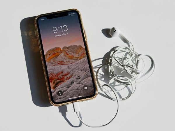 An iPhone on a white surface with white iPhone headphones connected to it, under a bright light shining from above that creates a sharp shadow under the phone and headphones.