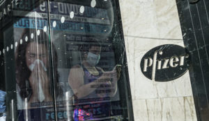 An ad for COVID-19 testing reflects on glass at a bus stop, as pedestrians walk past Pfizer world headquarters, Monday Nov. 9, 2020, in New York. (AP Photo/Bebeto Matthews)