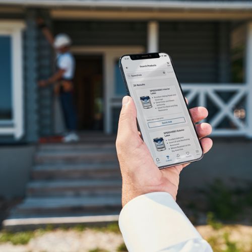 Person holding phone looking at PPG paint buckets with blurred front porch and person in background