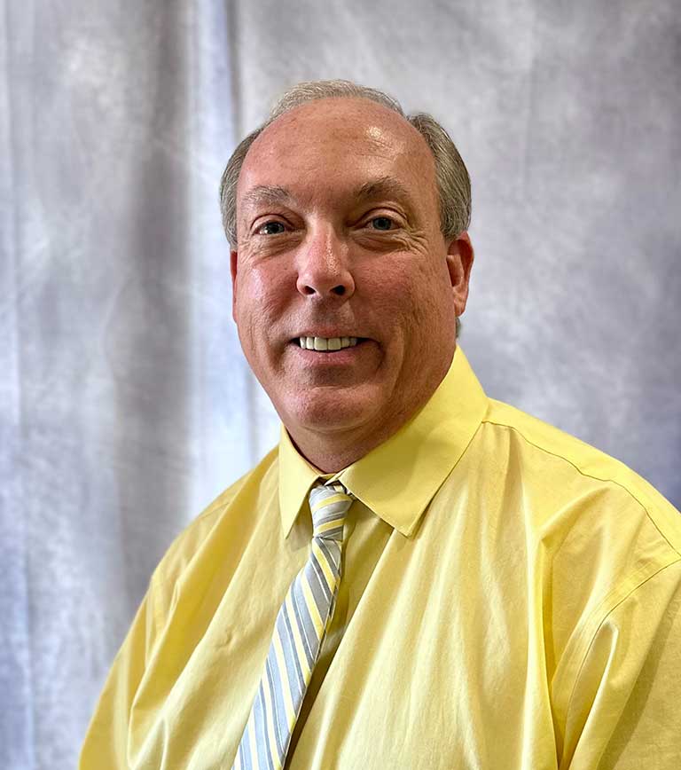 A man with short gray hair, wearing a yellow shirt and a striped tie, smiles at the camera in front of a gray backdrop.