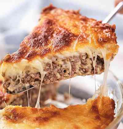 A close-up of a slice of cheesy meat pie being lifted from a baking dish, showing melted cheese and a browned top.