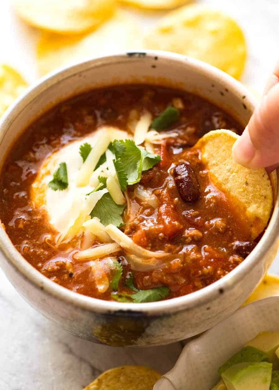 Corn chip being dipped into a bowl of Chili topped with sour cream, grated cheese and cilantro