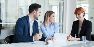 Three business people sit at a table and smile while looking at a tablet.