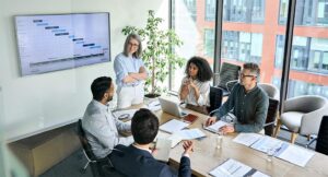 A woman gives a presentation in front of a boardroom table with other businesspeople.