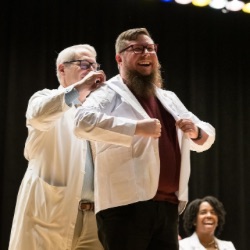 Two men in stand on stage, one helps the other put on a white coat, at the University of Rochester's white coat ceremony.