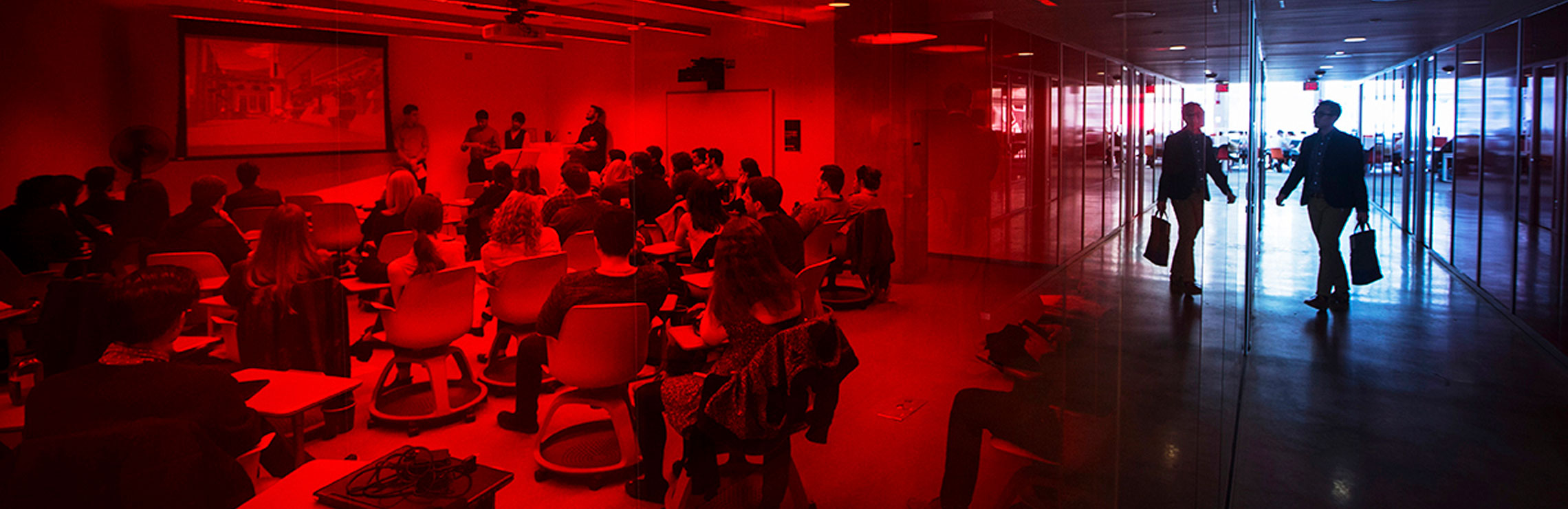 The sillouhette of a person walking down a hallway and entering a full classroom.