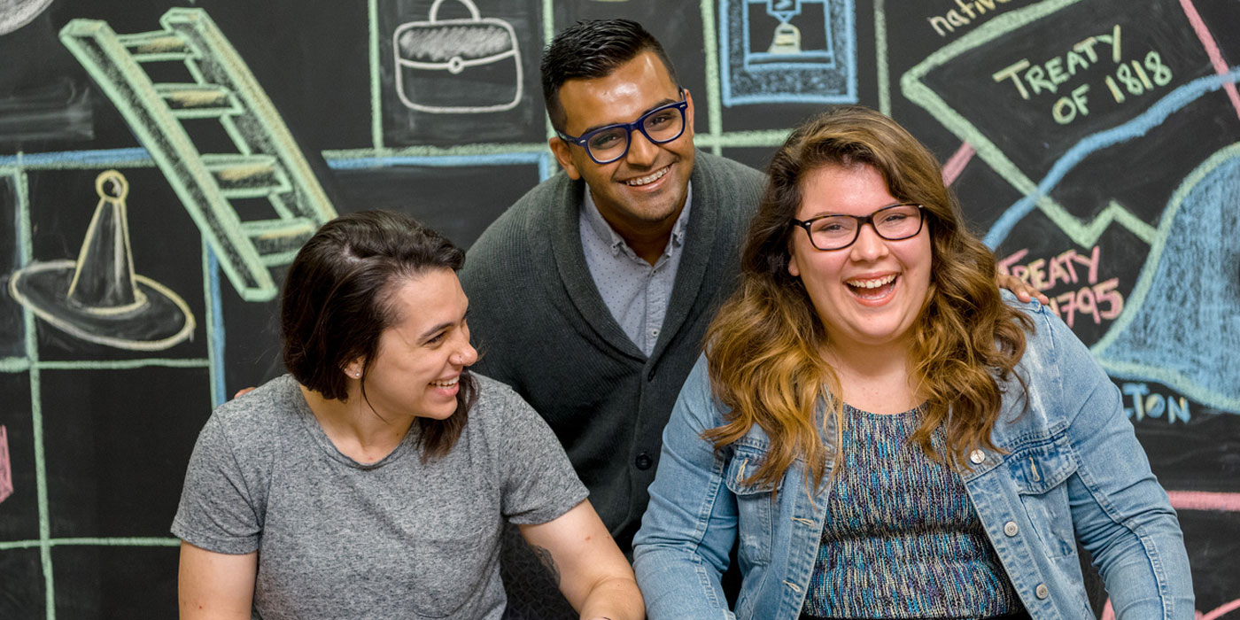 Three students candidly laughing while sitting in front of a colourful mural. 