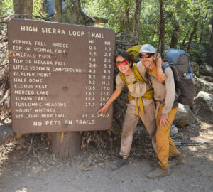 A teen boy and his father in muddy earth tone clothes, hiking backpacks, and sunglasses, smiling and pointing at a large park sign titled High Sierra Loop Trail. The teenager is pointing to the 211-mile long John Muir Trail route.