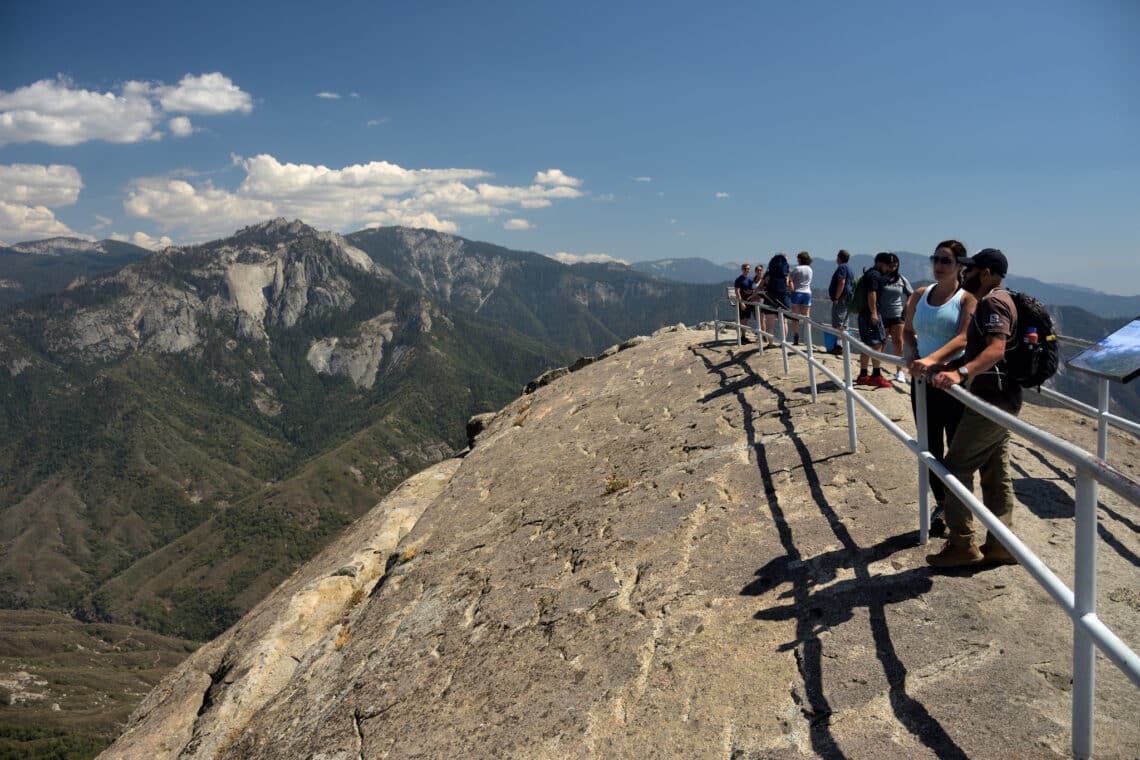 People stand behind a guardrail atop a rocky dome with mountains in the background.